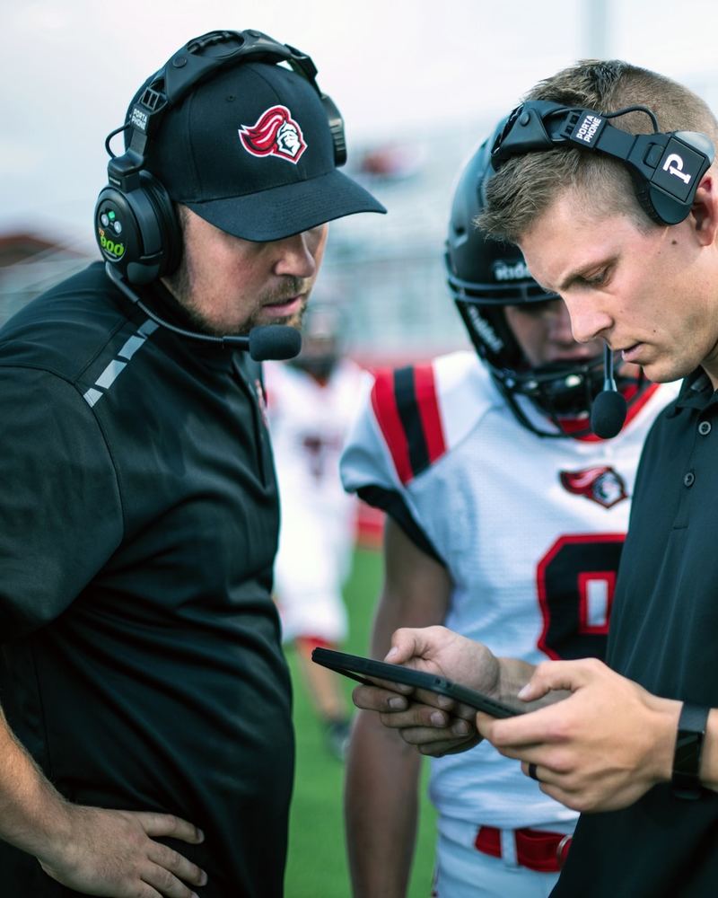Two football coaches in matching team polos and headsets on the sideline